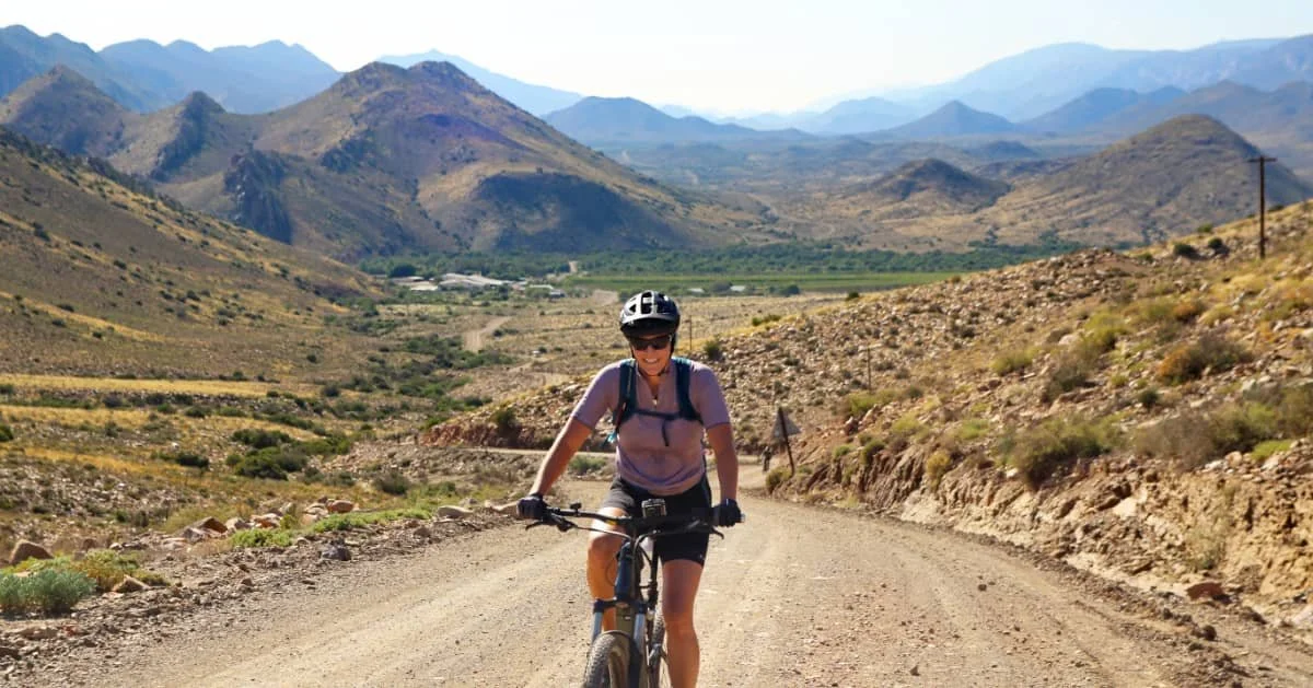 A woman riding a mountain bike on a dirt trail through a dry, hilly landscape with distant mountains.