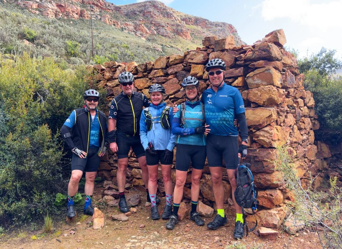 Five people in cycling gear standing together outdoors in front of a stone wall, with a mountain in the background.