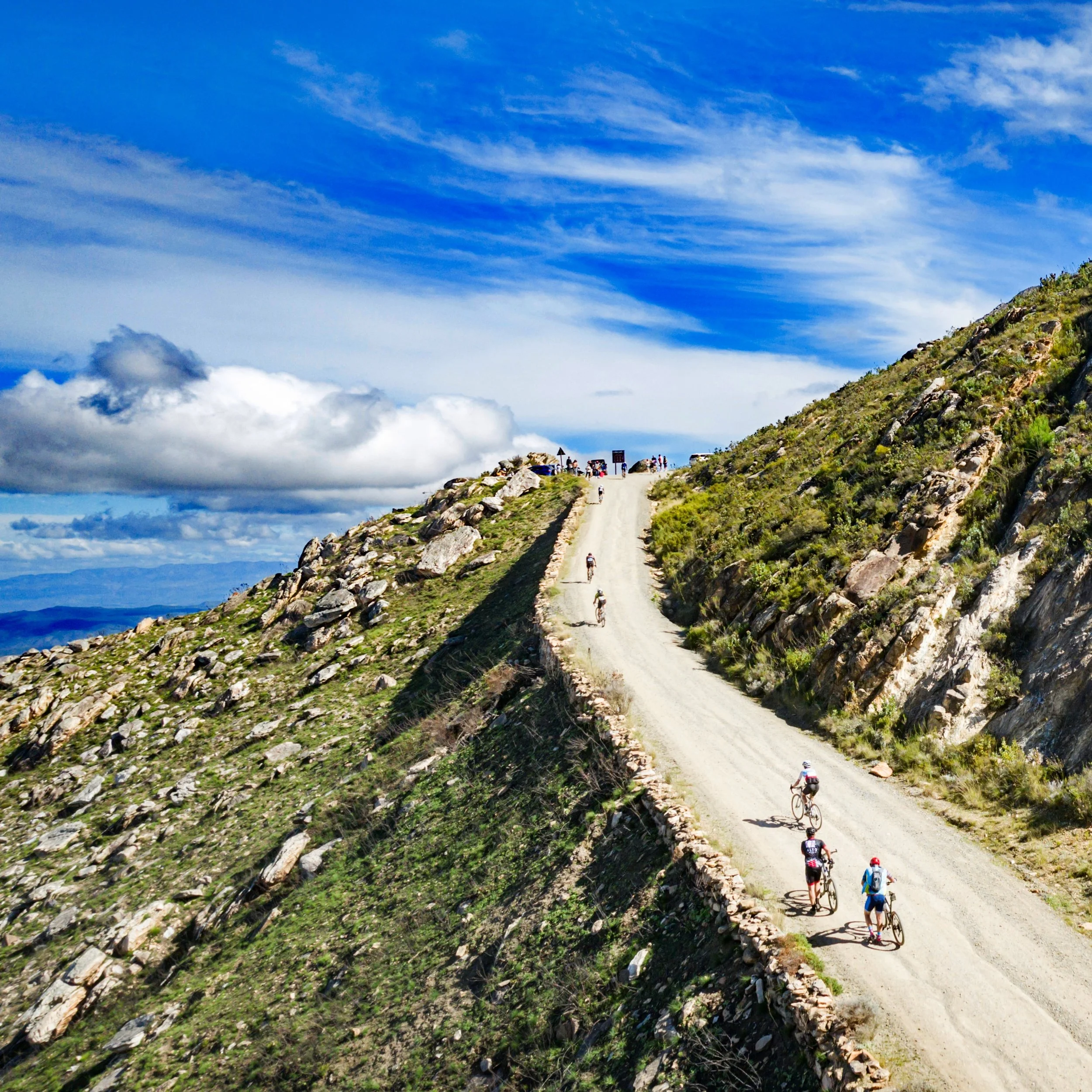 Cyclists riding on a mountain trail with a partly cloudy sky overhead and a scenic landscape.
