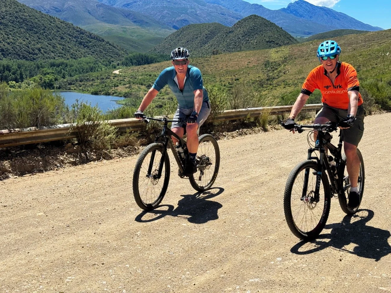 Two men riding mountain bikes on a dirt trail in a scenic mountainous area with a lake, green hills, and mountains in the background, under a clear blue sky.