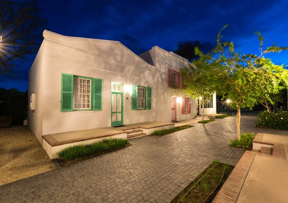 Nighttime view of a white house with teal and pink shutters, front door, and lit windows, surrounded by a paved courtyard, trees, and outdoor lights.