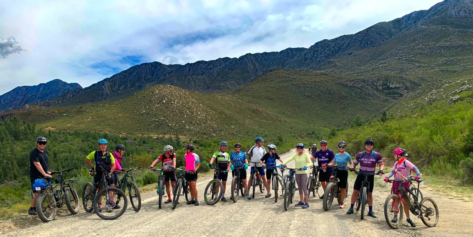 Group of 13 people with bicycles on a dirt road surrounded by green hills and mountains, under a partly cloudy sky.