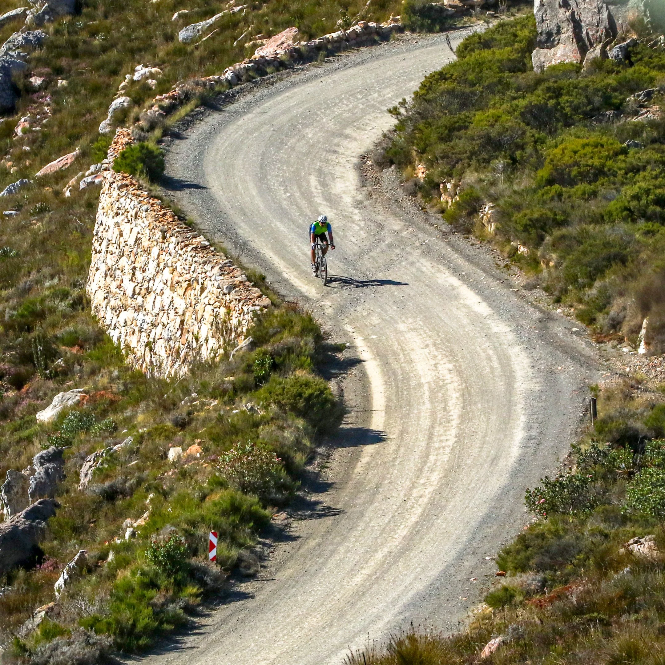 A person riding a bicycle on a winding dirt trail through a rocky, hilly landscape with green bushes and rocks.
