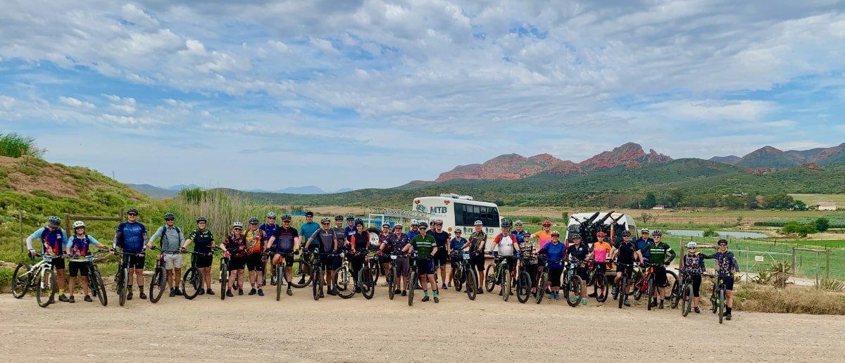 Group of people with bicycles at a desert trail, mountains, and cloudy sky in the background.