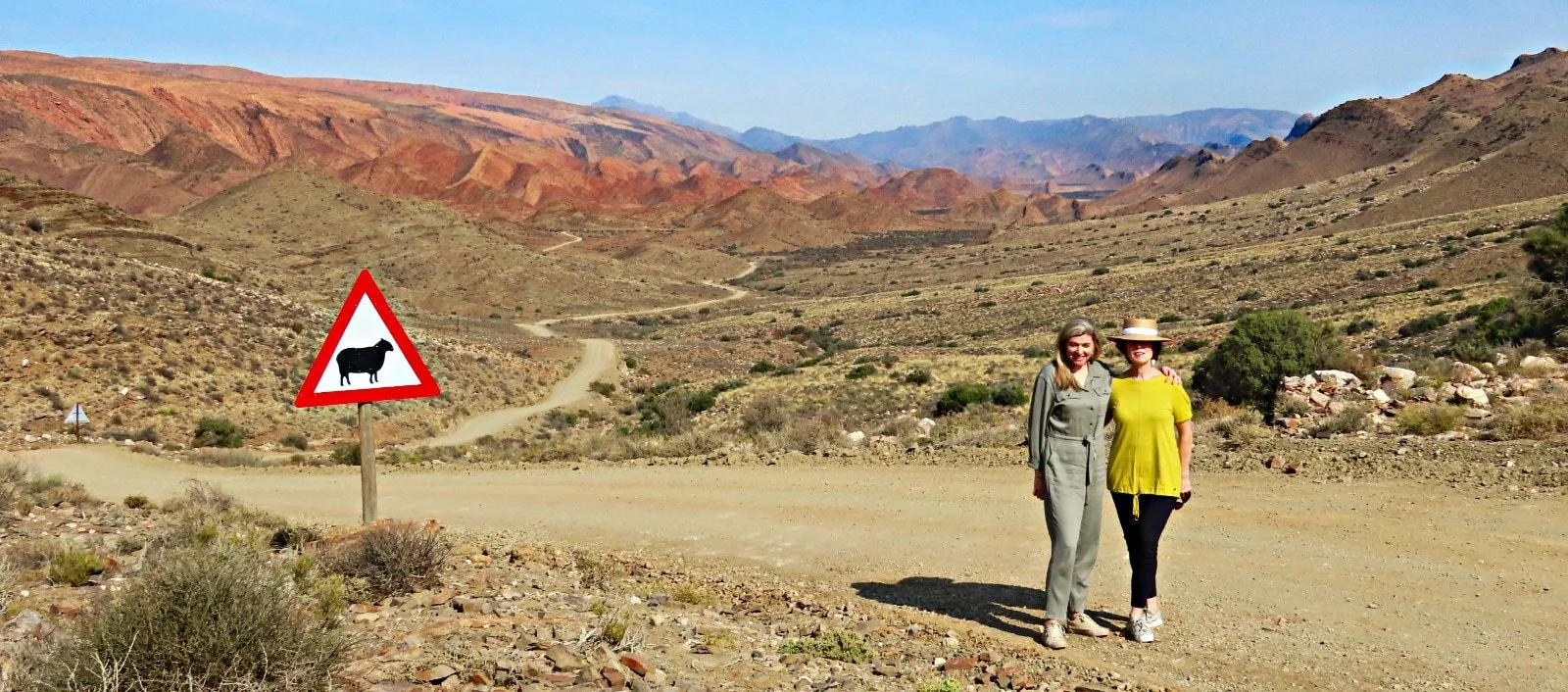 Two women standing on a dirt road in a desert landscape with mountains in the background, one wearing a green jumpsuit and the other in a yellow top with a large hat, near a warning sign indicating the presence of a sheep.