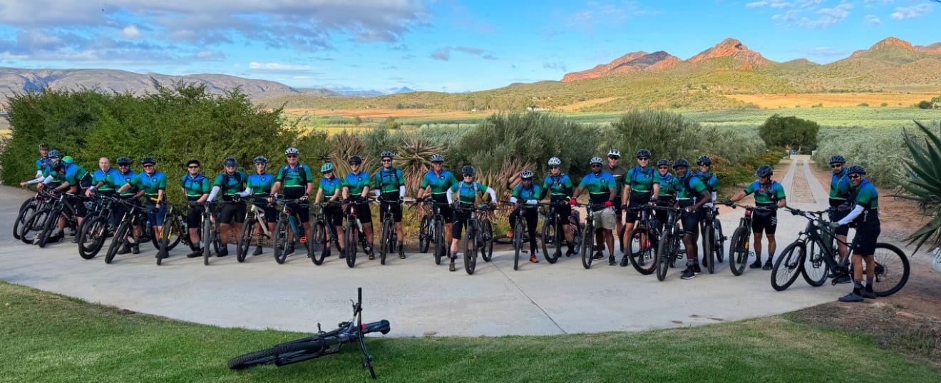 Group of cyclists in matching jerseys and helmets posing with bikes on a paved path in a scenic outdoor setting with mountains, trees, and clear sky in the background.