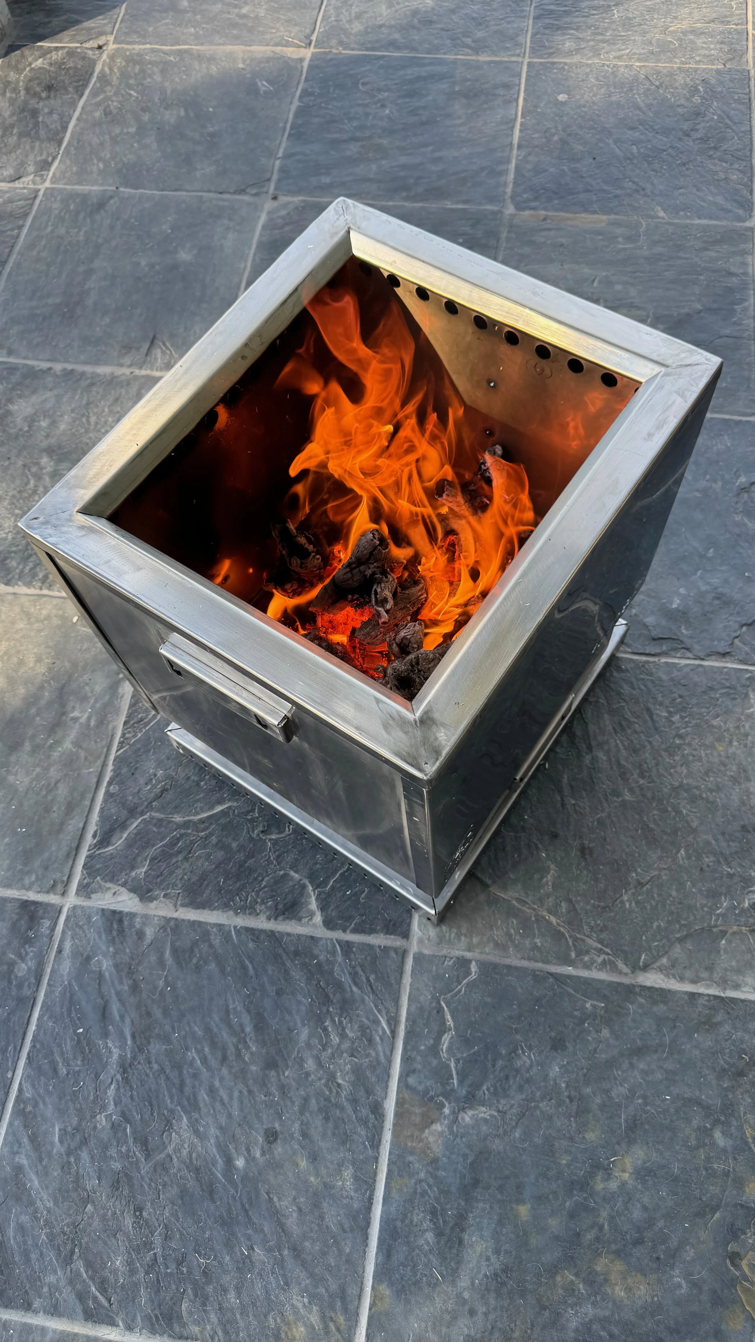 A portable stainless steel fire pit with burning wood and flames on a gray tiled outdoor surface.