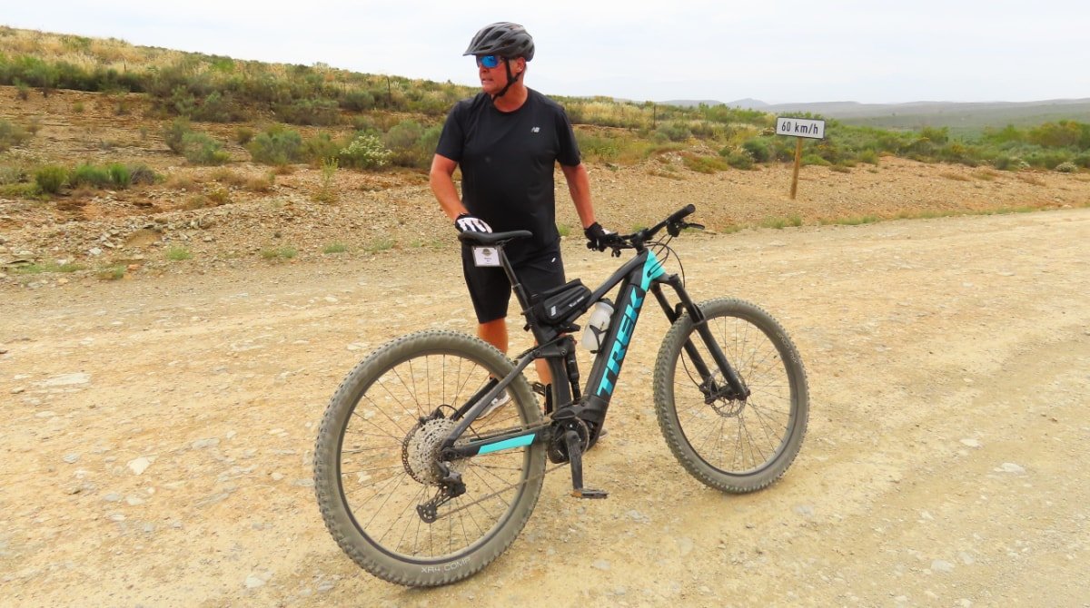 A person standing next to a black and teal TREK mountain bike on a dirt trail, wearing a helmet, sunglasses, and athletic clothes, with a landscape of dry, rocky terrain and sparse vegetation in the background.