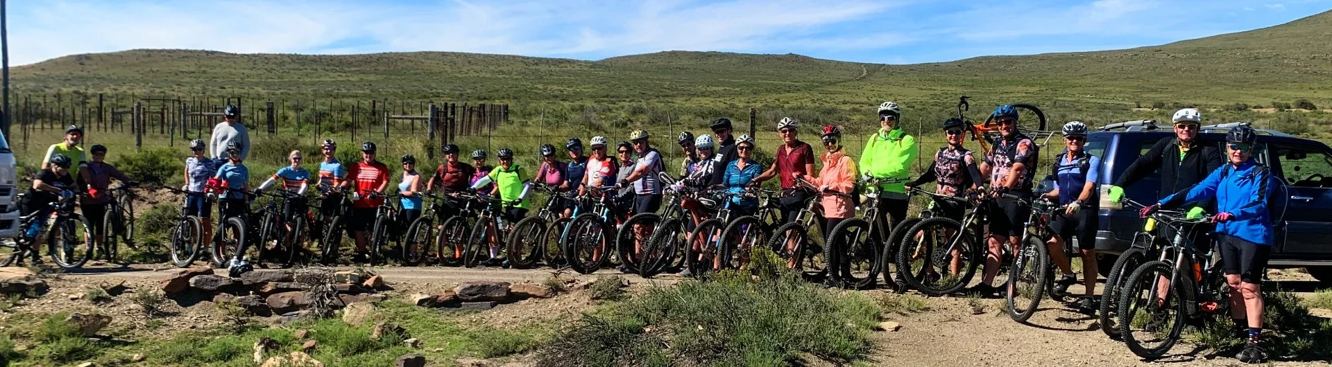 A large group of people with bicycles posing for a photo outdoors near a desert trail with hills in the background.
