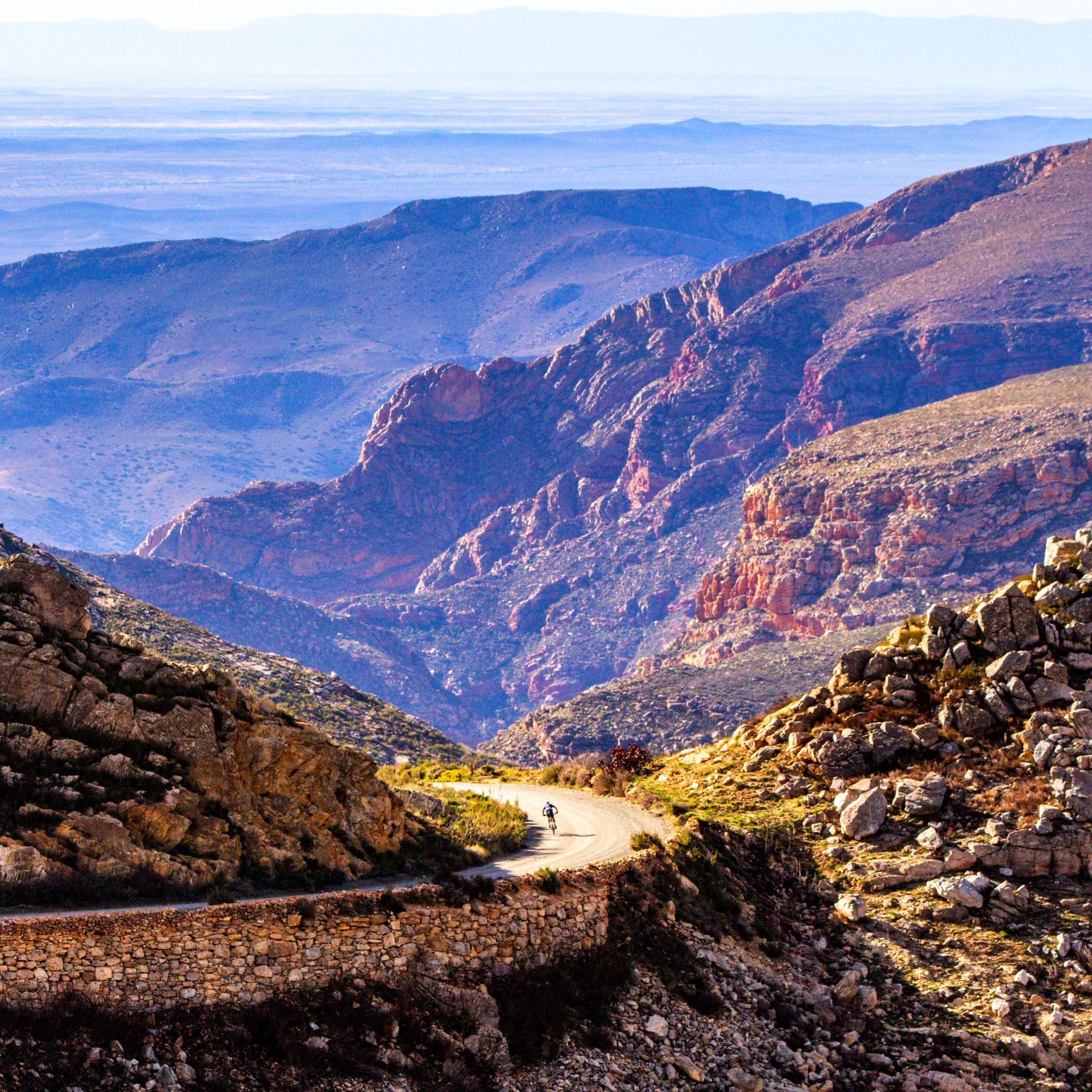 A mountain landscape with colorful rocky slopes and a winding dirt road with a lone cyclist.