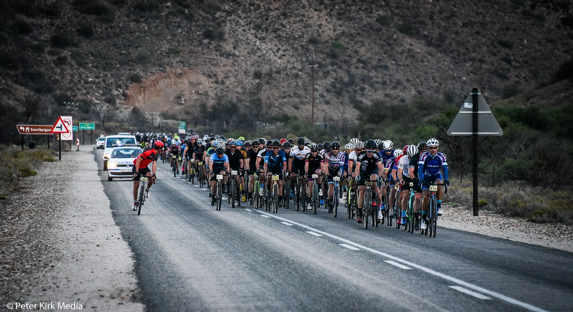 A large group of cyclists ride on a mountain road with police cars leading and following behind. The background features arid hills and sparse vegetation.