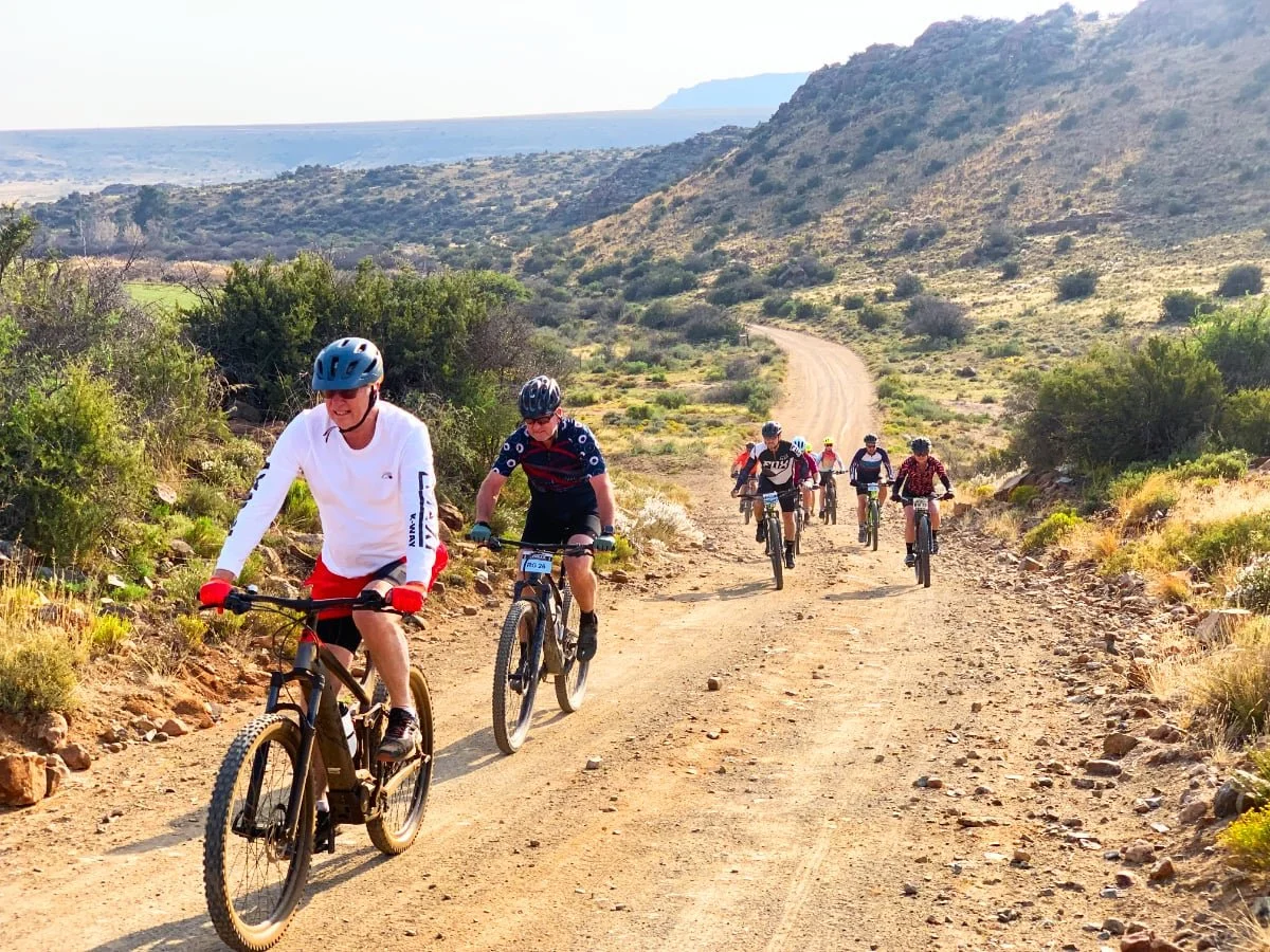 Group of people mountain biking on a dirt trail in a desert landscape with hills and sparse vegetation.