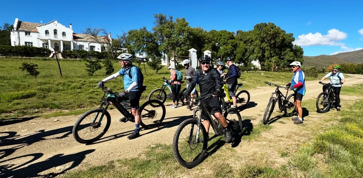 Group of people on mountain bikes riding on a dirt trail in a rural area with green grass, trees, and a white building in the background under a blue sky.