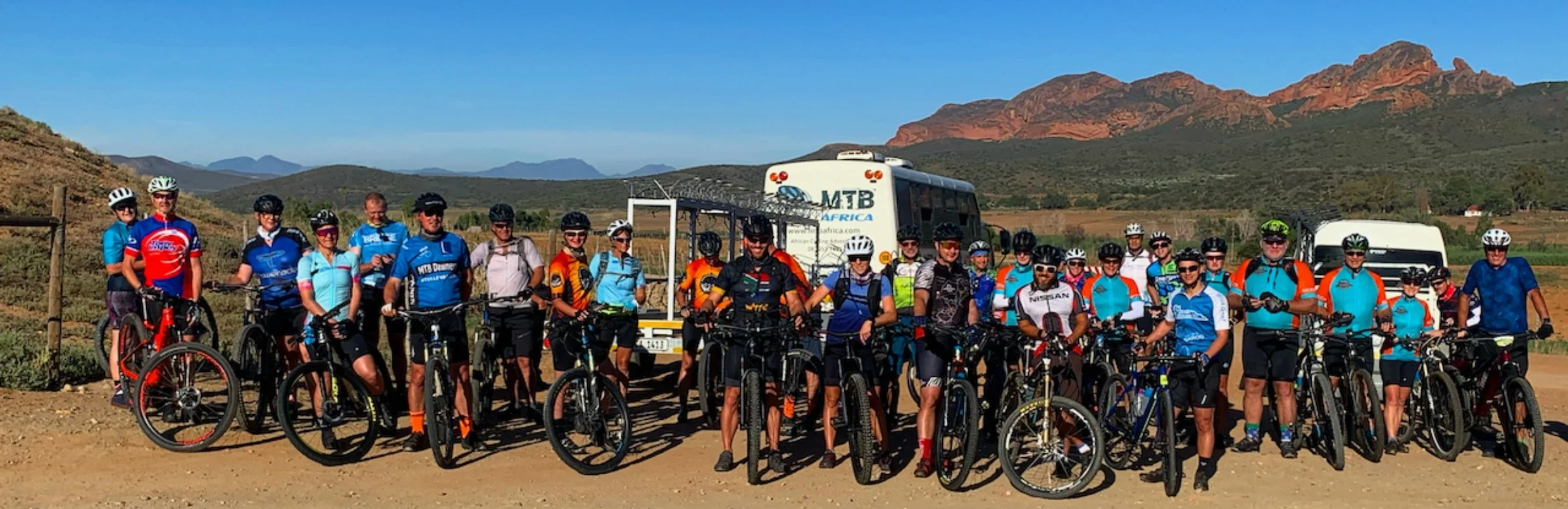A group of cyclists gathered for a photo outdoors with mountains in the background, standing beside their bikes and two support vehicles.