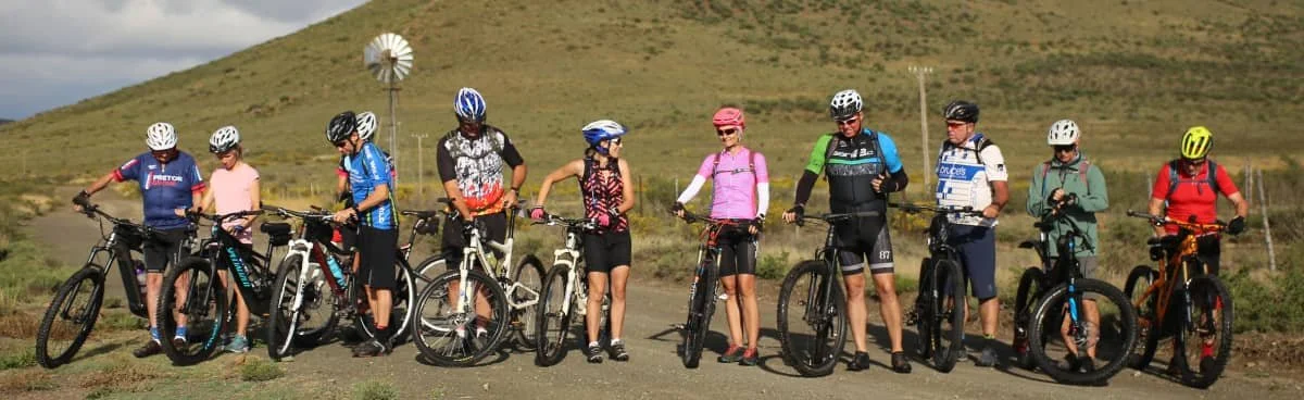 Ten people appear to be getting ready for a bike ride on a dirt trail. They are holding or standing next to their mountain bikes, wearing helmets and casual clothing, with a rural landscape and hills in the background.