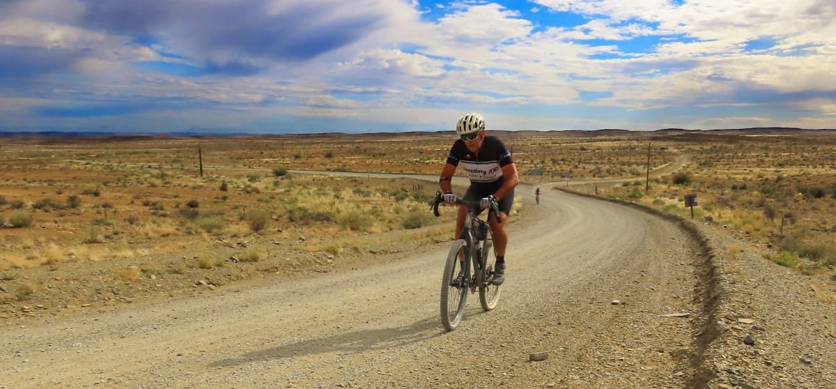 A man riding a mountain bike on a dirt road in a desert landscape under a cloudy sky.