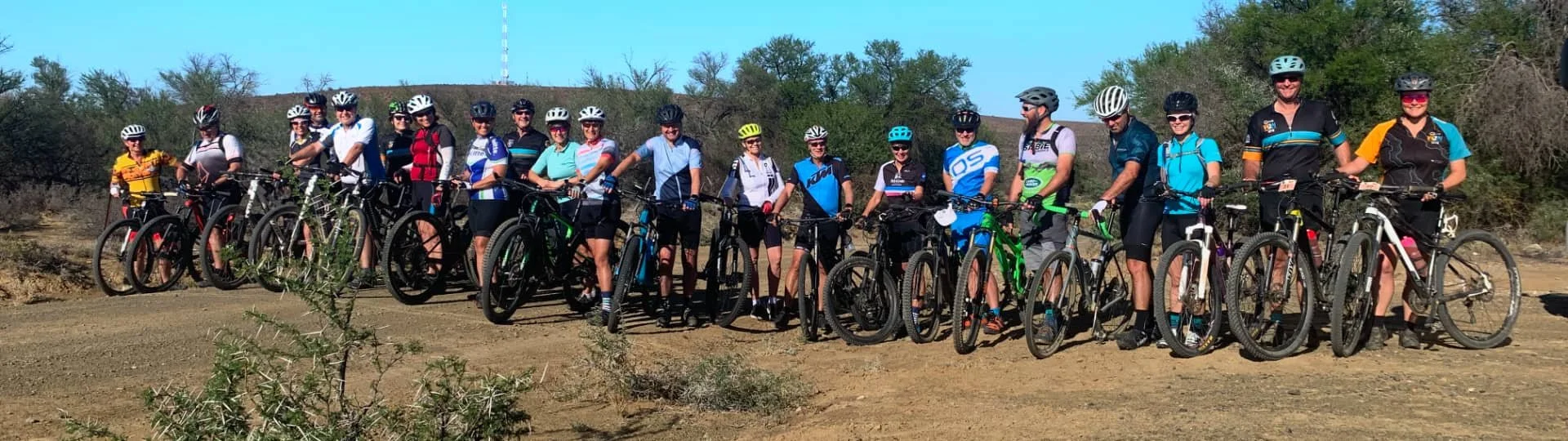 Group of people with bicycles standing outdoors on a dirt path, wearing helmets, in a desert-like area with dry bushes and trees in the background.