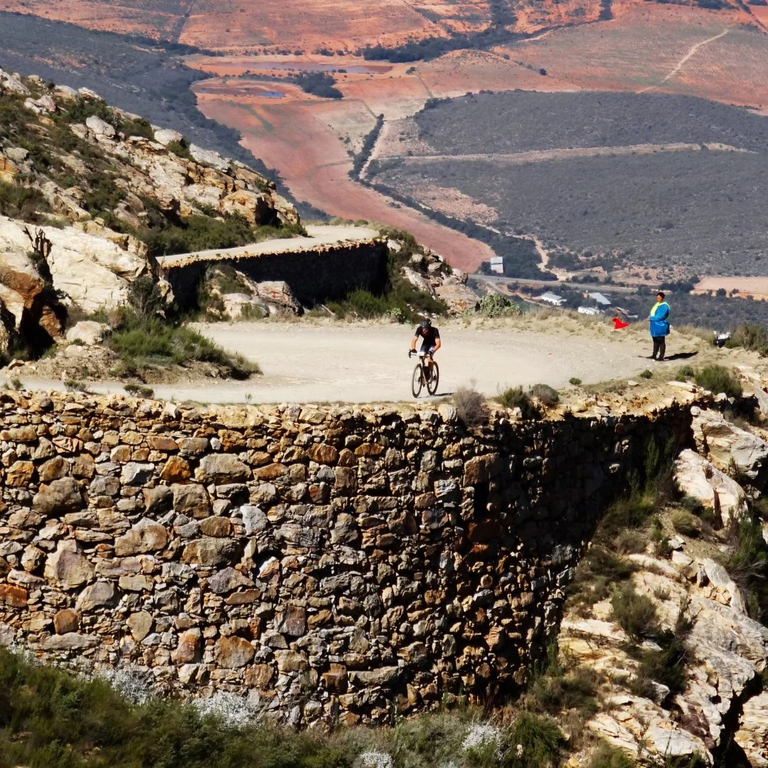 A cyclist riding on a winding mountain road with a stone retaining wall, surrounded by rocky terrain and sparse vegetation, with a person standing nearby holding a red flag, and a landscape of rolling hills in the background.