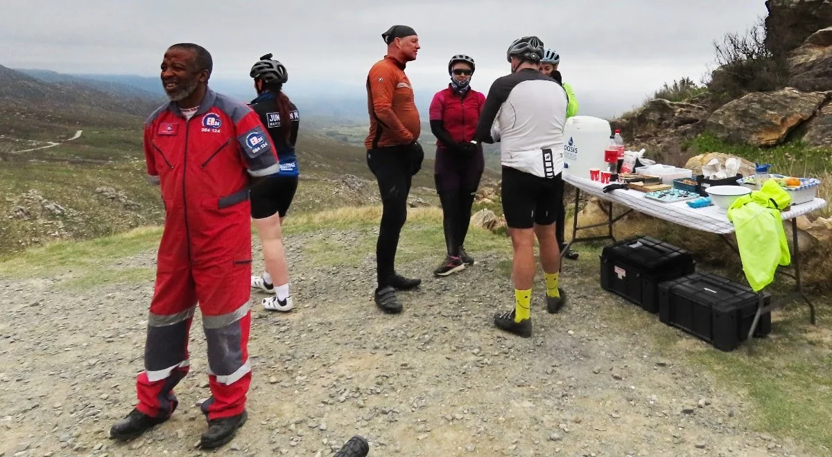 A group of people in cycling gear standing near a table with food and drinks on a mountain trail overcast sky
