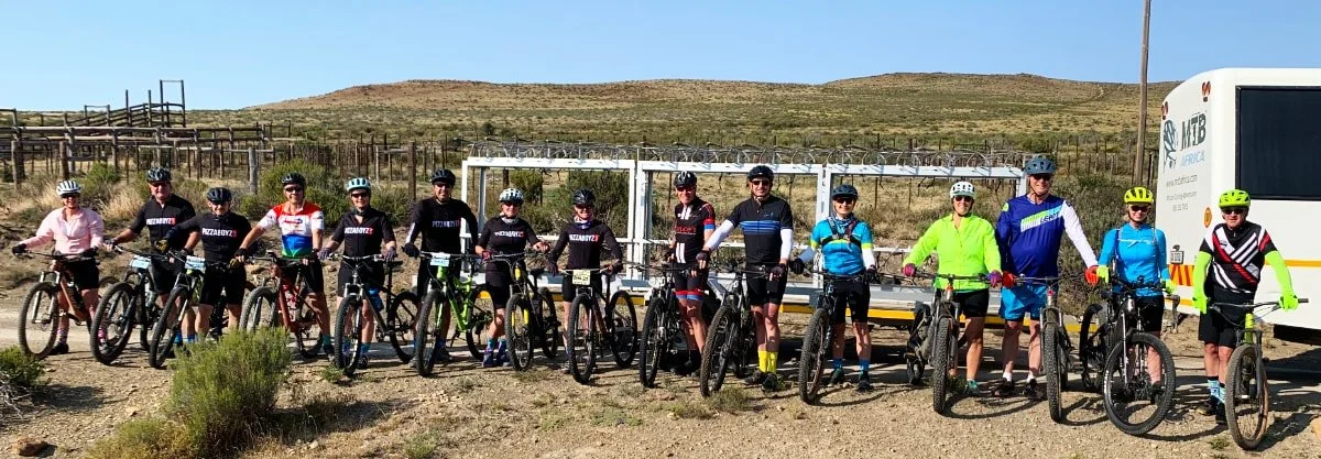 Group of cyclists with gear, standing with their bikes outdoors in a desert landscape, some wearing helmets and colorful clothing, with a trailer and vehicle in the background.