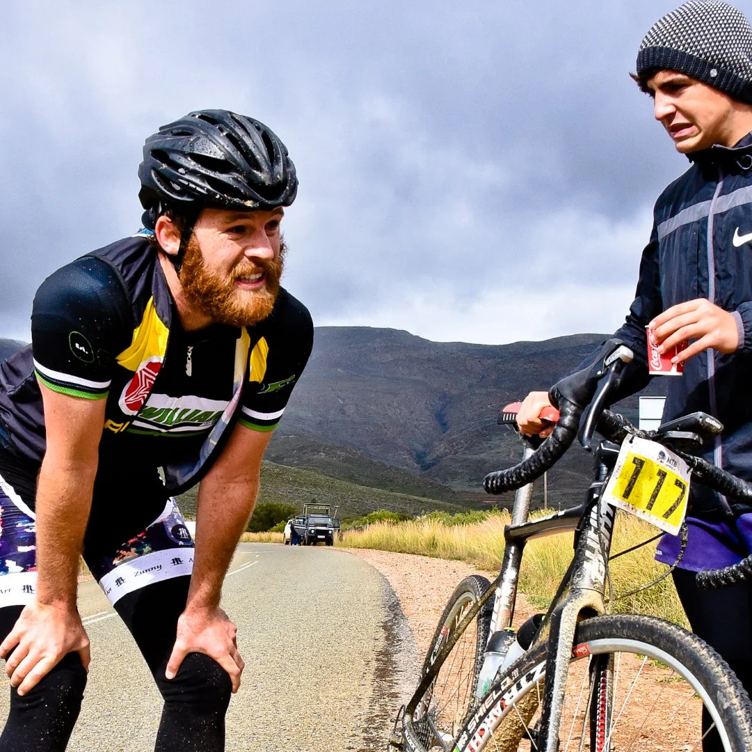 A man with a bicycle and a race number talking to another man on a mountain road with hills in the background.