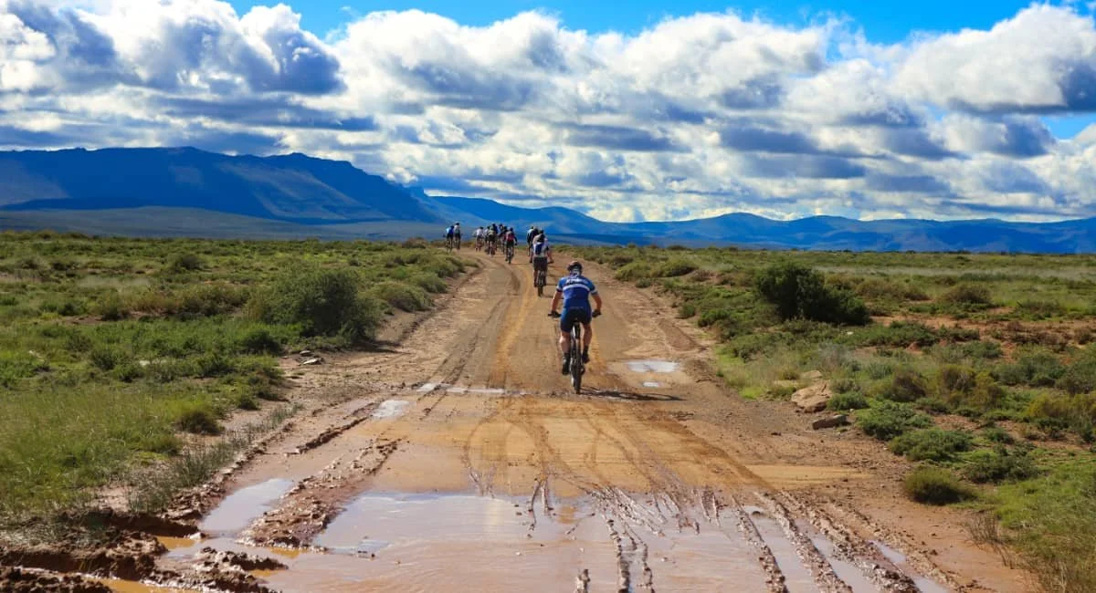 A group of cyclists riding on a muddy dirt trail through a grassy plain with mountains and a partly cloudy sky in the background.