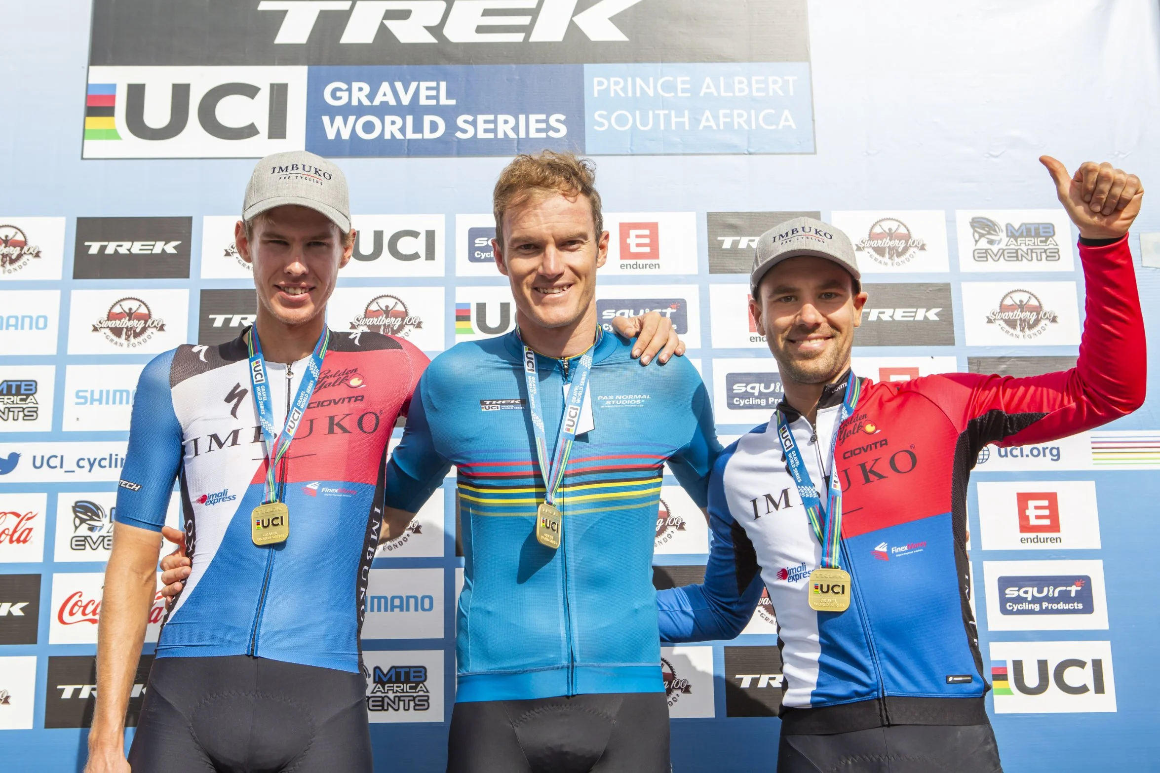 Three male cyclists stand on a winners' podium holding medals. The cyclist in the middle is smiling and has his arm around the other two. They are wearing cycling uniforms and caps. Behind them is a backdrop displaying logos and text related to the U