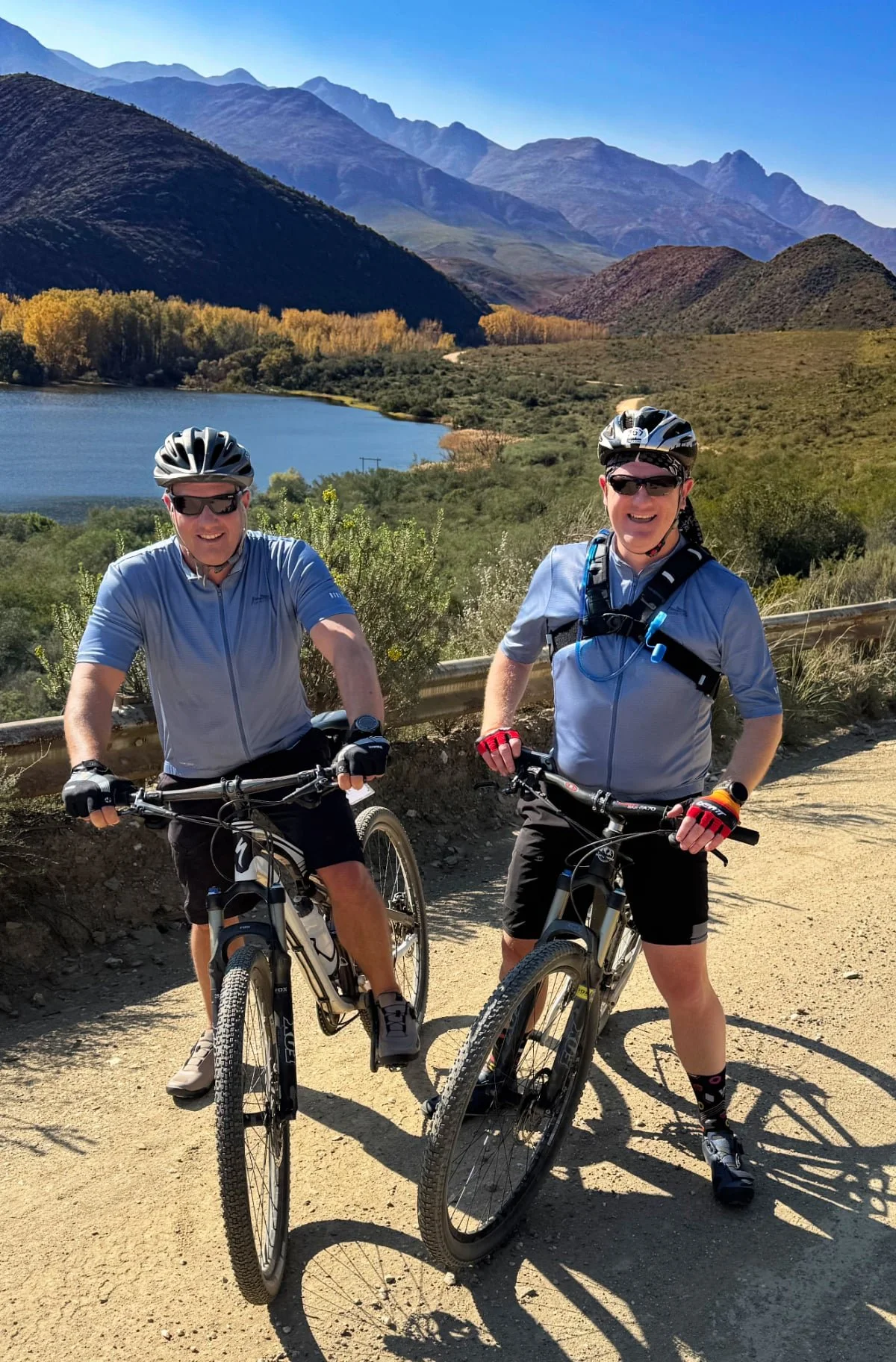 Two men in cycling gear posing with mountain bikes on a dirt trail with a scenic landscape of mountains, trees, water, and a clear blue sky in the background.