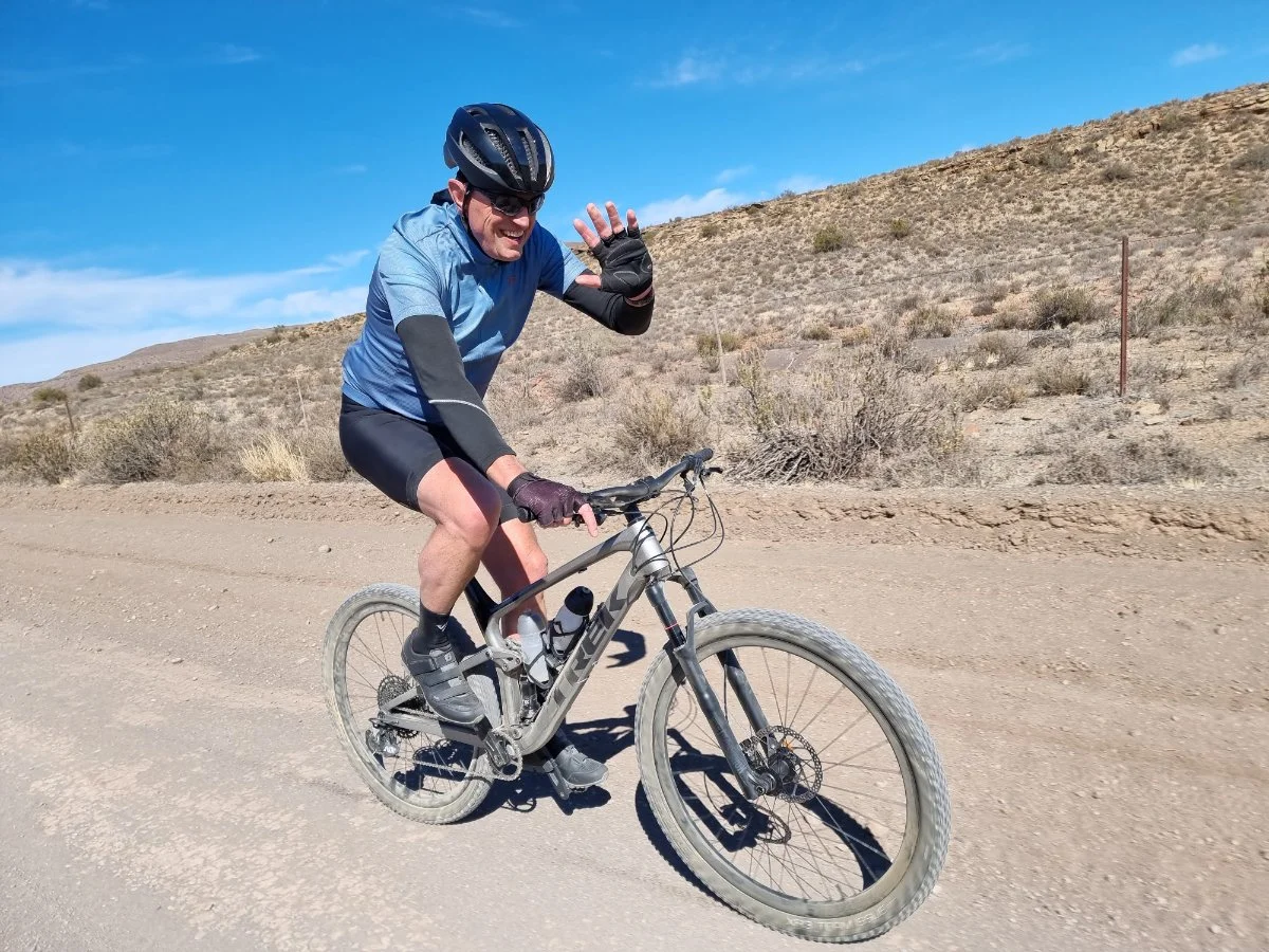 A man riding a mountain bike on a dirt trail in a desert landscape, wearing a helmet, sunglasses, and sportswear, waving and smiling.