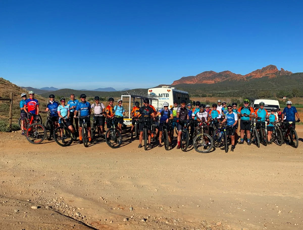 A large group of cyclists posing with their bikes outdoors in a scenic area with mountains in the background, under a clear blue sky.