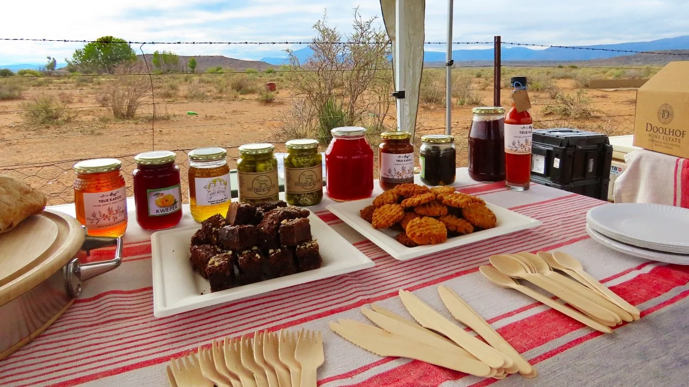 A table set outdoors in a desert landscape with jars of honey, jars of preserves, and plates of baked goods and fried chicken, under a tent with a mountain view in the background.