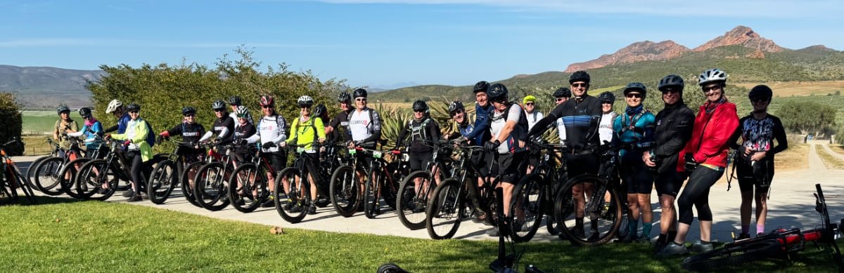 Group of cyclists lined up outdoors with mountains and blue sky in the background.