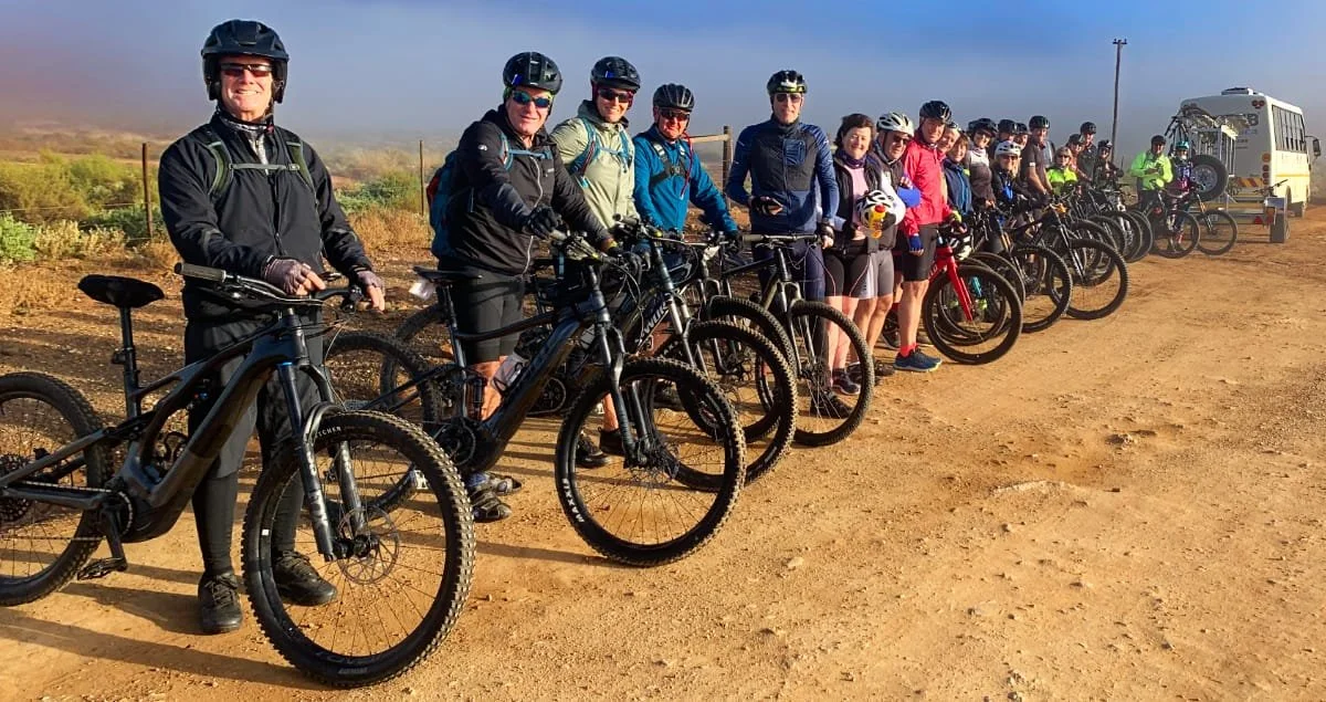 A group of people in cycling gear with helmets standing with mountain bikes on a dirt trail, with a bus and power poles in the background.