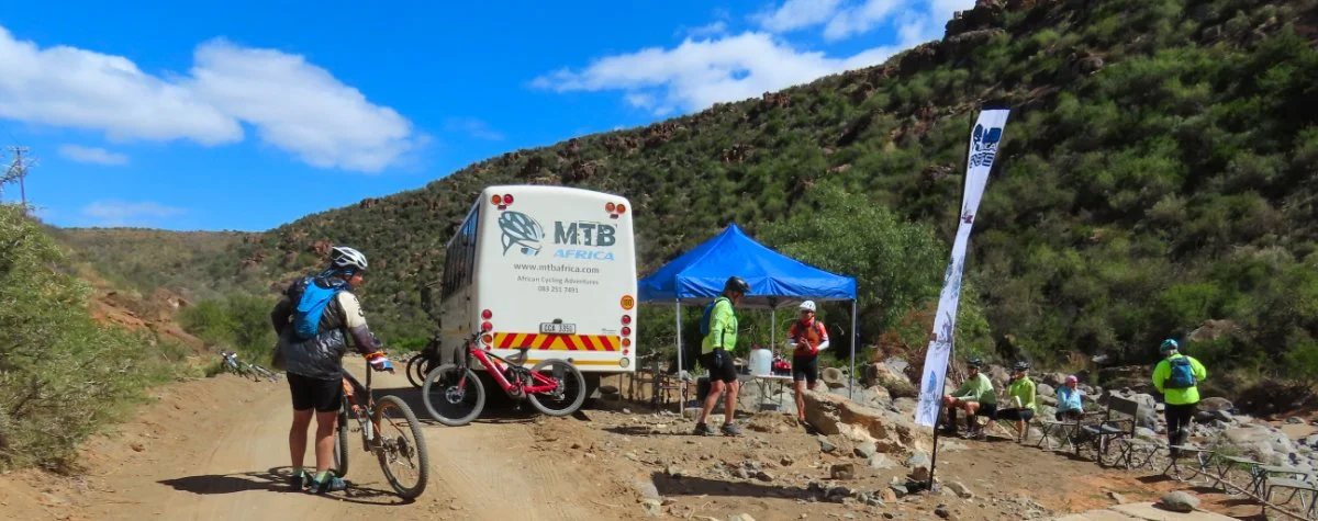 Mountain bikers resting at a trailhead with a blue canopy tent, a support vehicle, and a flag, surrounded by hills and rocky terrain.