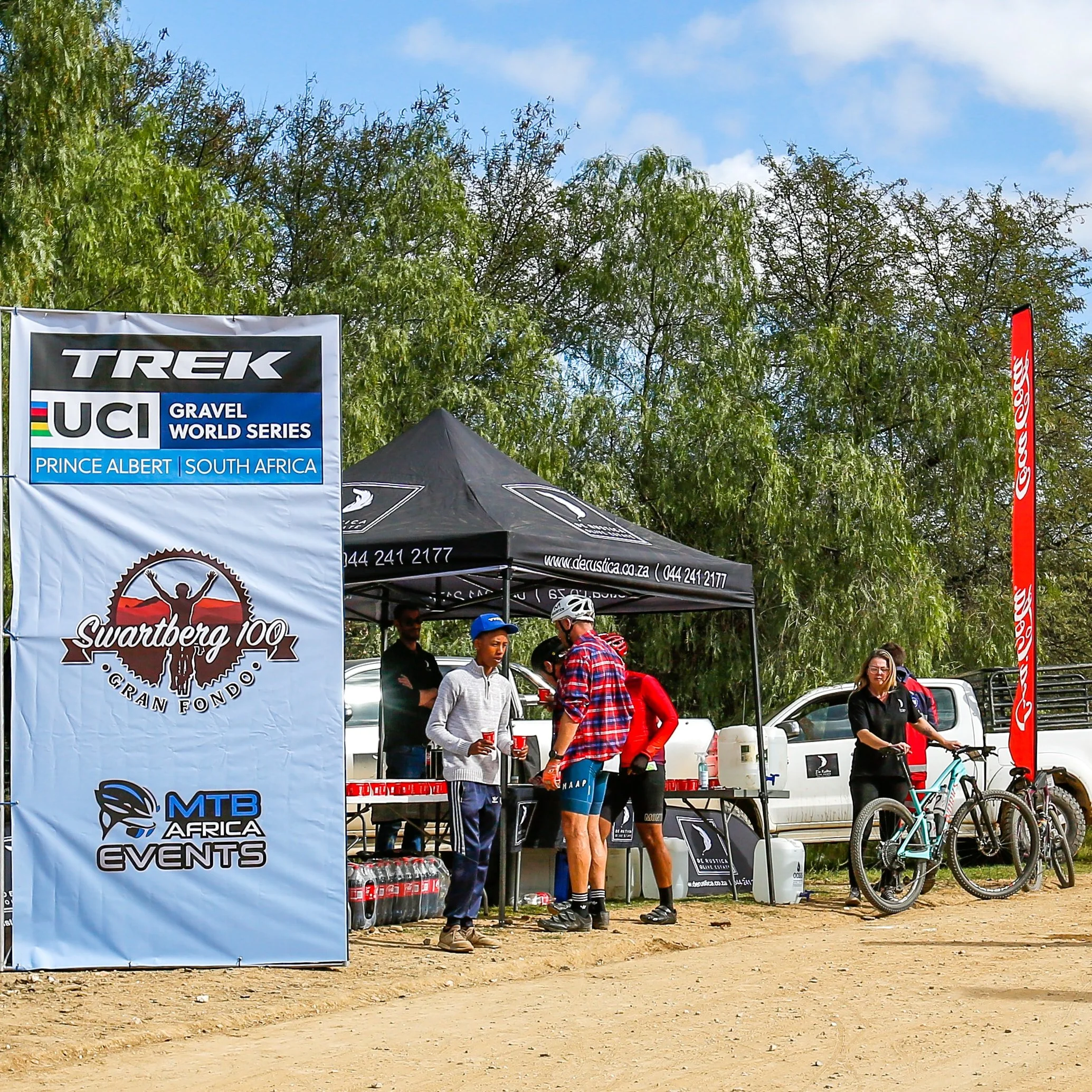 People gathered at a mountain biking event setup area with banners, tents, and bicycles, outdoors on a dirt path with trees and a blue sky in the background.