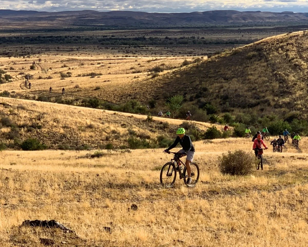 A group of people riding mountain bikes along a dirt trail through a dry, grassy landscape with hills and mountains in the distance.