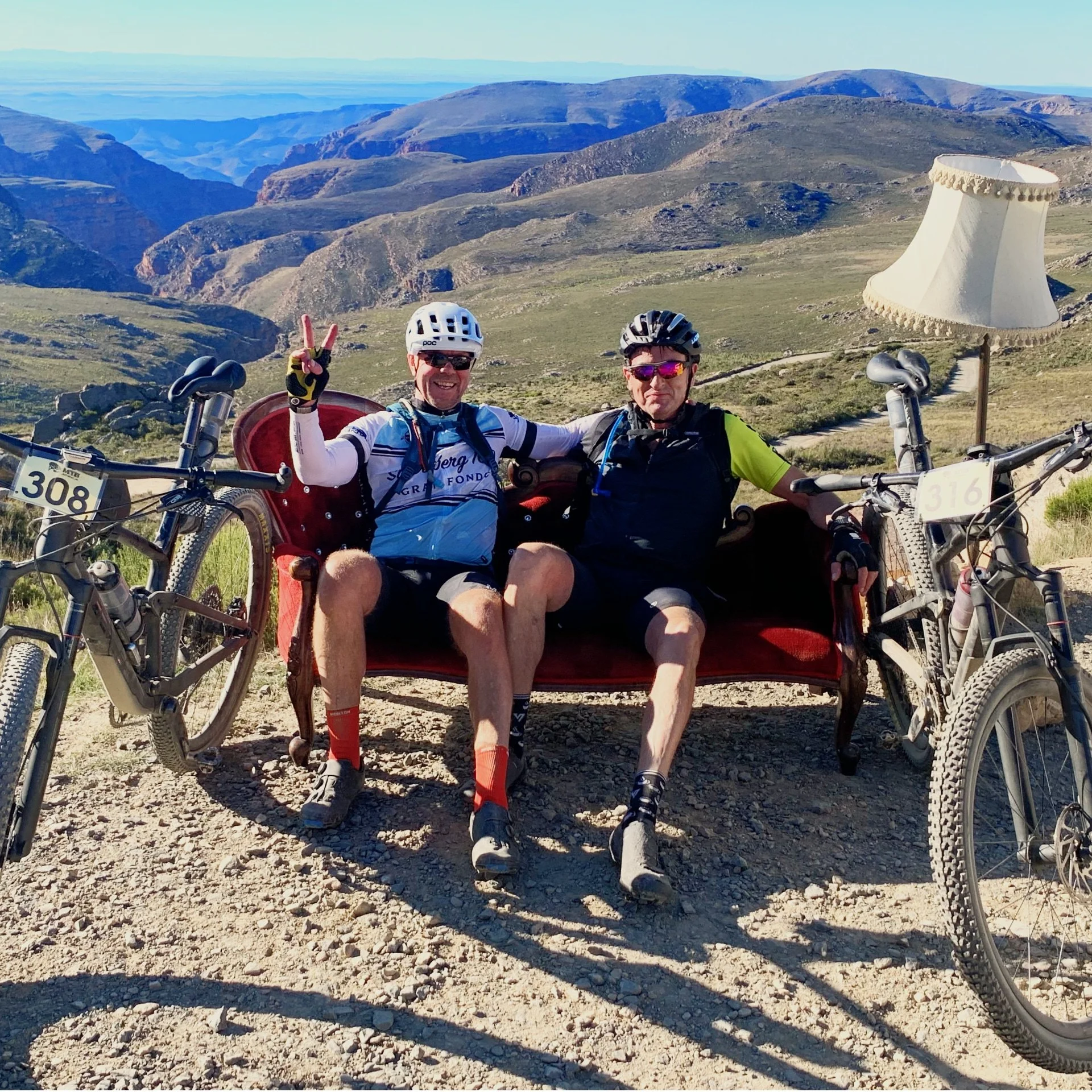Two cyclists taking a break on a rocky trail in a mountainous landscape, sitting on a vintage red sofa with a lampshade nearby, with bicycles leaning on either side.