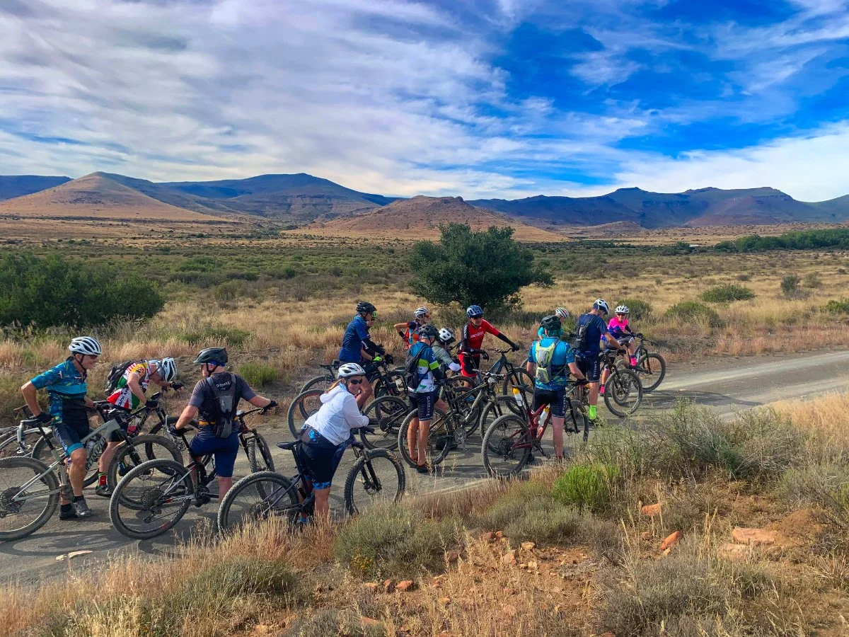 Group of cyclists resting on a trail in a desert landscape with mountains and a partly cloudy sky in the background.