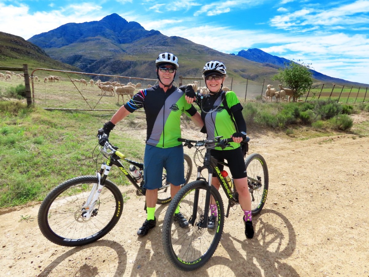 Two cyclists, a man and a woman, posing with their bikes in a rural landscape with mountains, a fenced pasture, sheep, and a tree in the background.