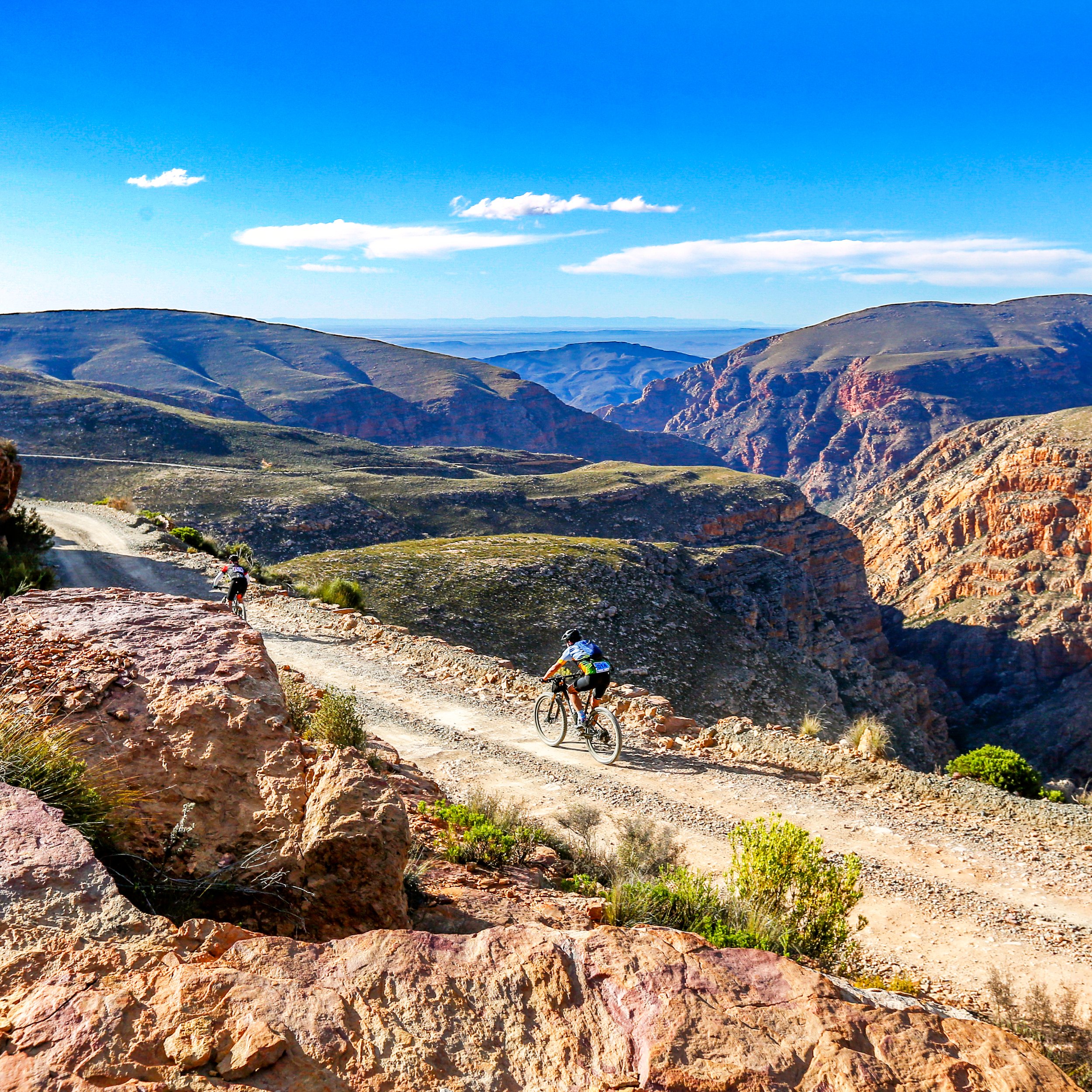 Two mountain bikers riding on a rugged dirt trail through a mountainous landscape with colorful cliffs, green vegetation, and a clear blue sky.