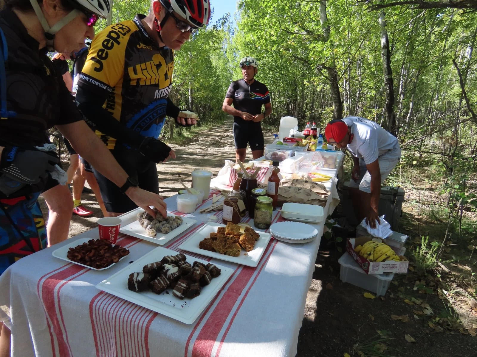 Group of cyclists at a refreshment table on a forest trail, with food and drinks.
