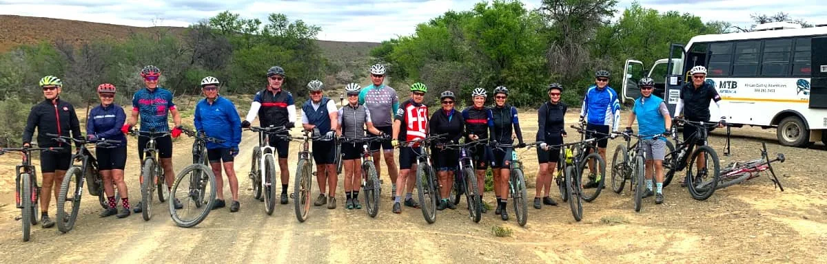 Group of 14 cyclists with bikes standing in a line on a dirt path near a bus, with trees and hills in the background.