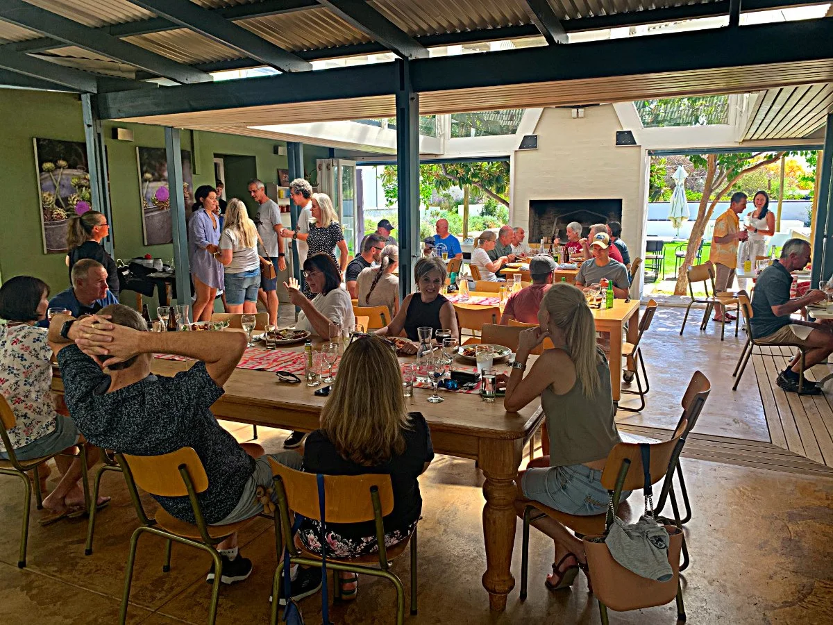 People dining and socializing at a restaurant with outdoor seating, green walls, and large windows showing greenery outside.