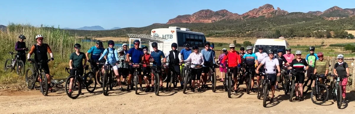 A group of people with bicycles on a dirt road, wearing helmets, in a rural area with mountains and blue skies in the background.
