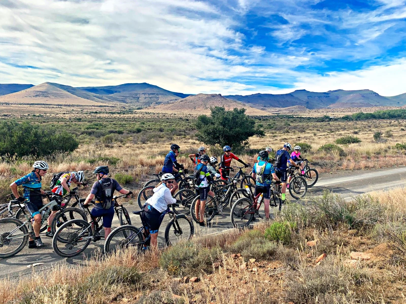 A group of cyclists taking a break on a dirt road in a desert landscape with mountains and a partly cloudy sky in the background.