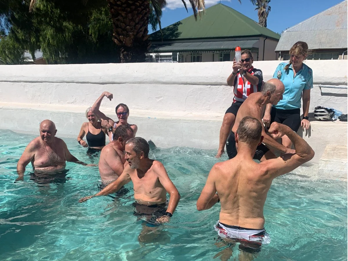 A group of seniors enjoying a swim in an indoor pool with some people flexing and others standing or sitting around the pool edge, under bright sunlight.
