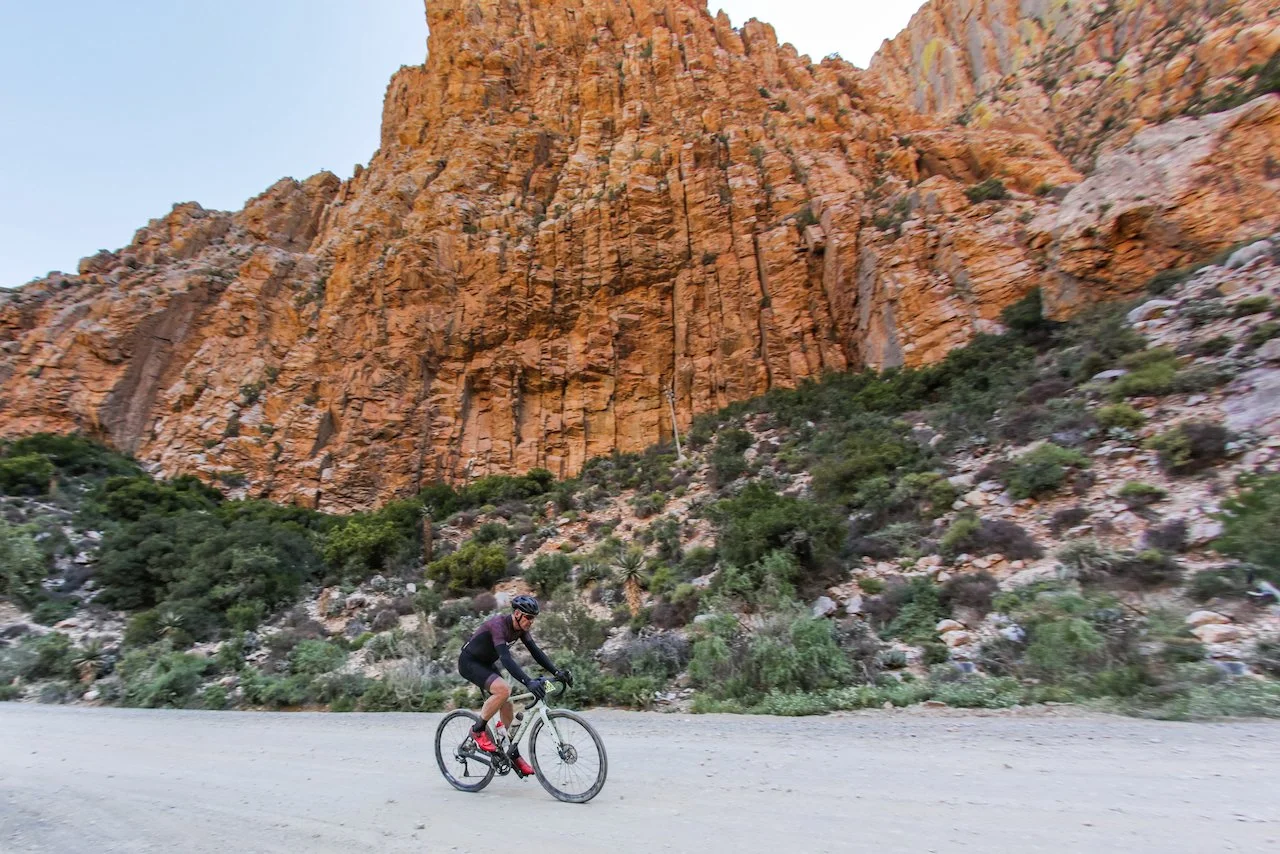 A person mountain biking on a trail with a large rocky orange cliff and green bushes in the background.
