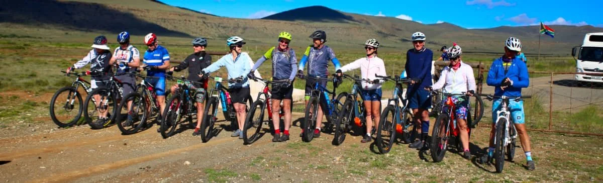 Group of people with mountain bikes standing outdoors on a dirt path with a mountain range in the background.