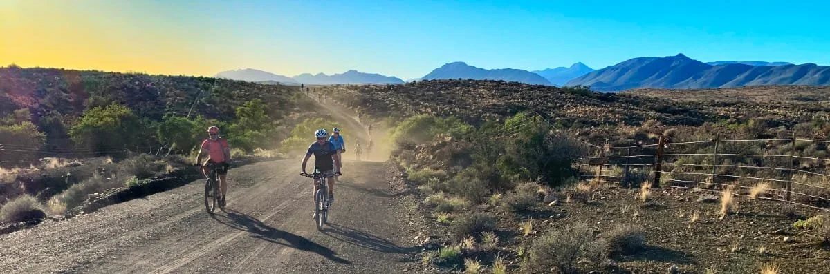 Three cyclists riding on a dirt trail in a desert landscape with mountains in the background under a clear blue sky.