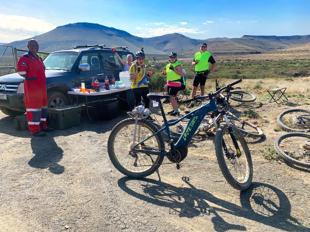 Group of people resting during a mountain biking trip in a desert landscape with mountains in the background. There are bikes on the ground, a table with drinks, and an emergency vehicle.